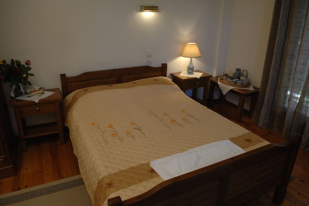A neatly made bed with a quilt, a bedside lamp, a nightstand with a book and a vase of flowers, and a small table with a tray of tea cups.