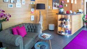 A modern reception area with a grey sofa, a small round table with magazines, a reception desk with a lamp, and framed pictures on the wall. - Villa Verde (Salzburg)