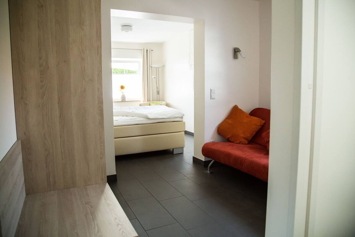 A modern hotel room with a bed, a red sofa, and a view of the outdoors.
