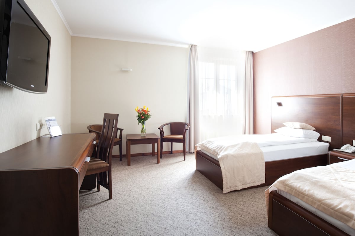 A hotel room with a desk, two chairs, a small table with a vase, a bed with a headboard, and a television mounted on the wall.