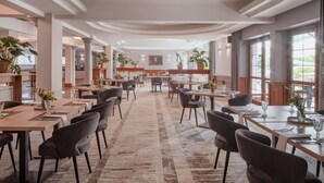 A spacious dining area with multiple tables and chairs, large windows, and potted plants. - Artis Hotel & Spa (Zamosc)
