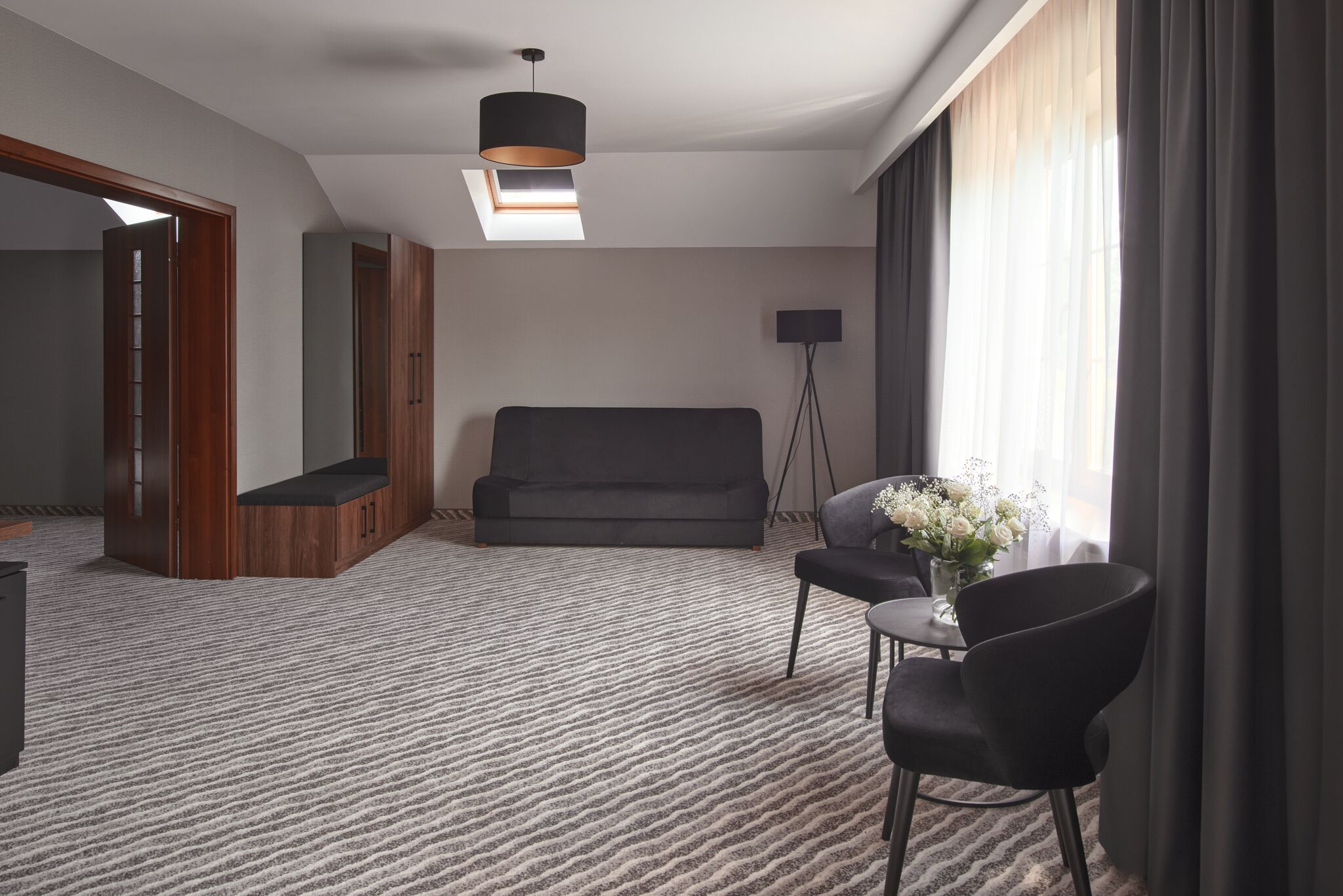 A modern hotel room with a large window, a sofa, a small table with a vase of flowers, and a floor lamp.