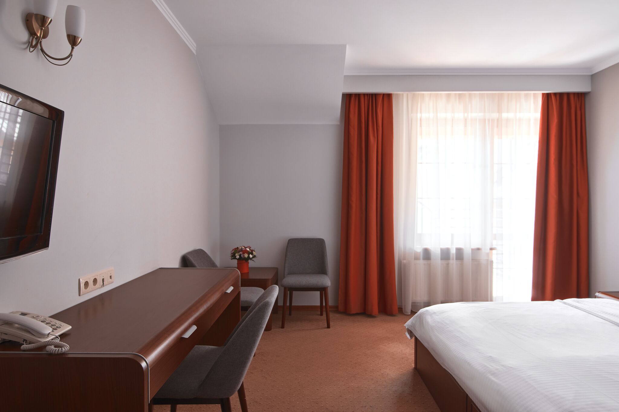 A hotel room with a bed, a desk with a telephone, a chair, a small table with a flower vase, and a television mounted on the wall.
