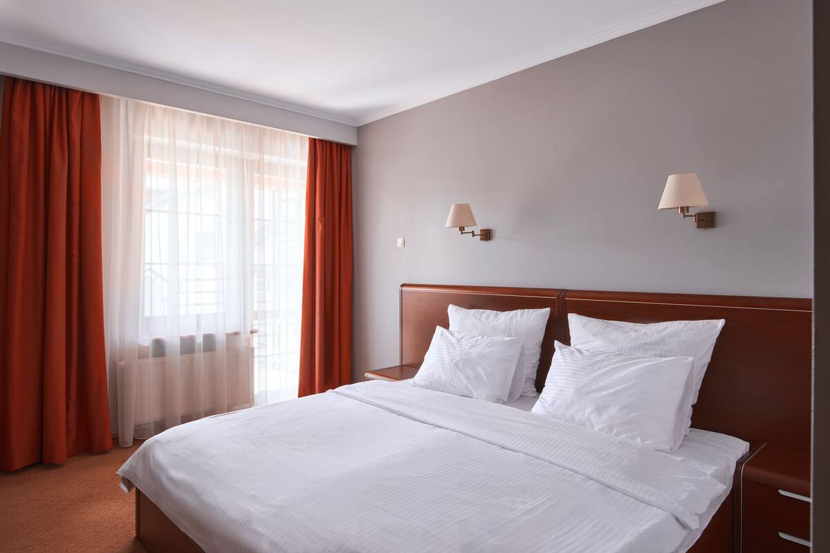 A hotel room with a large bed, two wall-mounted lamps, and a window with sheer curtains.