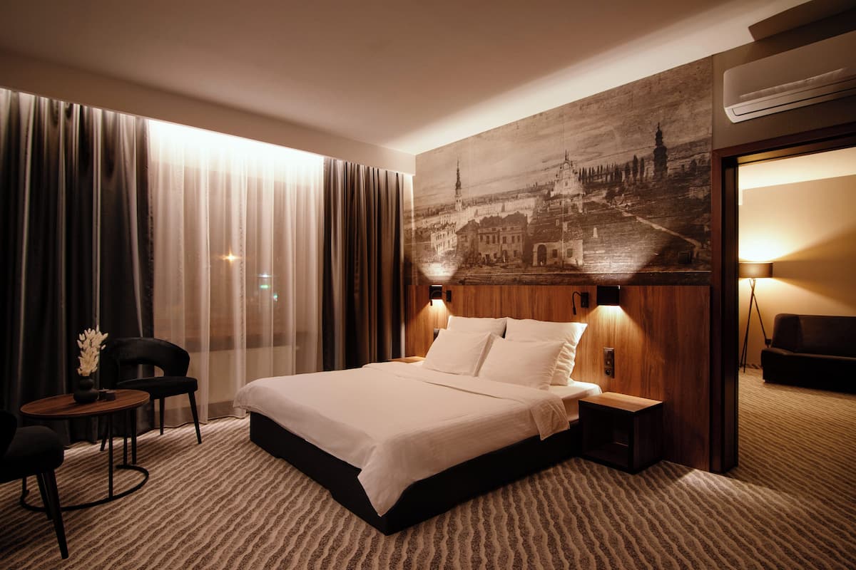A hotel room with a large bed, a small table, a chair, and a wall-mounted lamp.