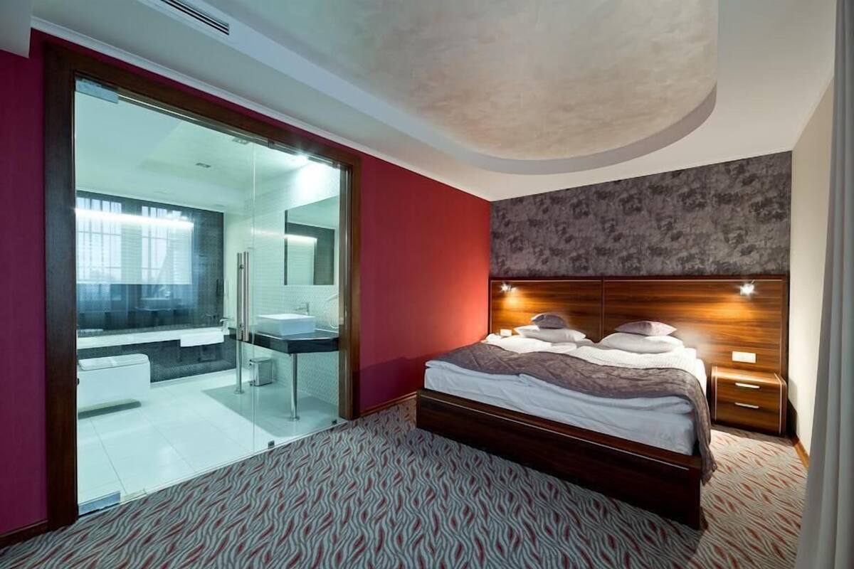 A modern hotel room with a glass-enclosed bathroom, a large bed with a wooden headboard, and a patterned carpet.
