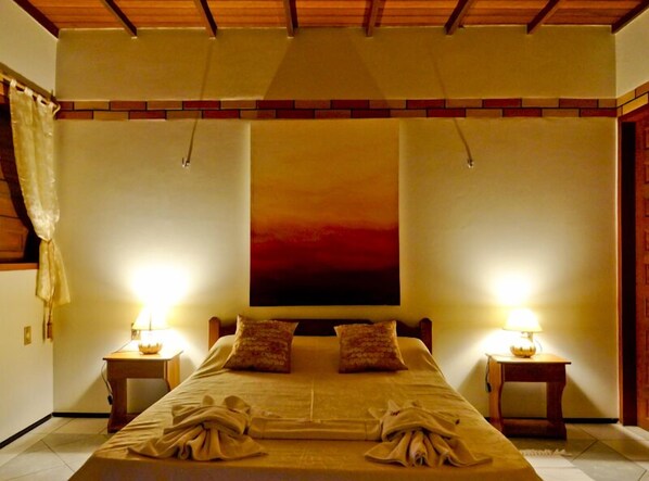 Ein Hotelzimmer mit einem großen Bett, zwei Nachttischen mit Lampen, einer Holzbalkendecke und einem Gemälde eines roten Sonnenuntergangs an der Wand.
