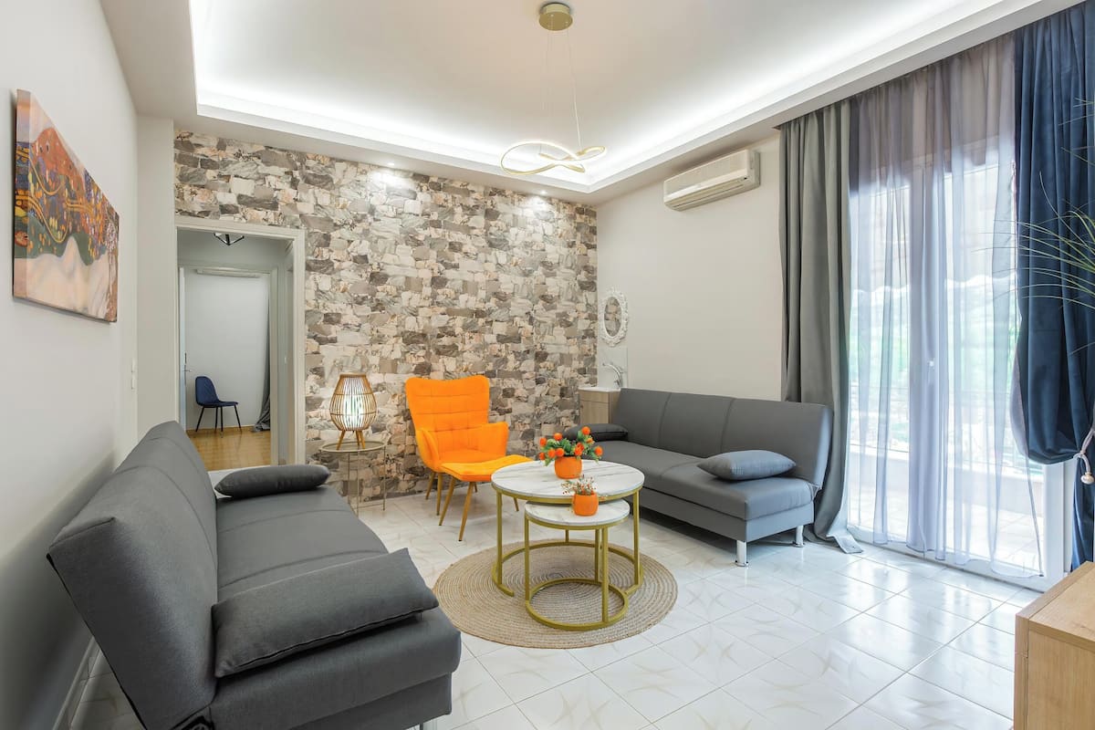 Una sala de estar moderna con pared de piedra, un sofá gris, un sillón naranja, una mesa de centro redonda y un ventanal con cortinas.