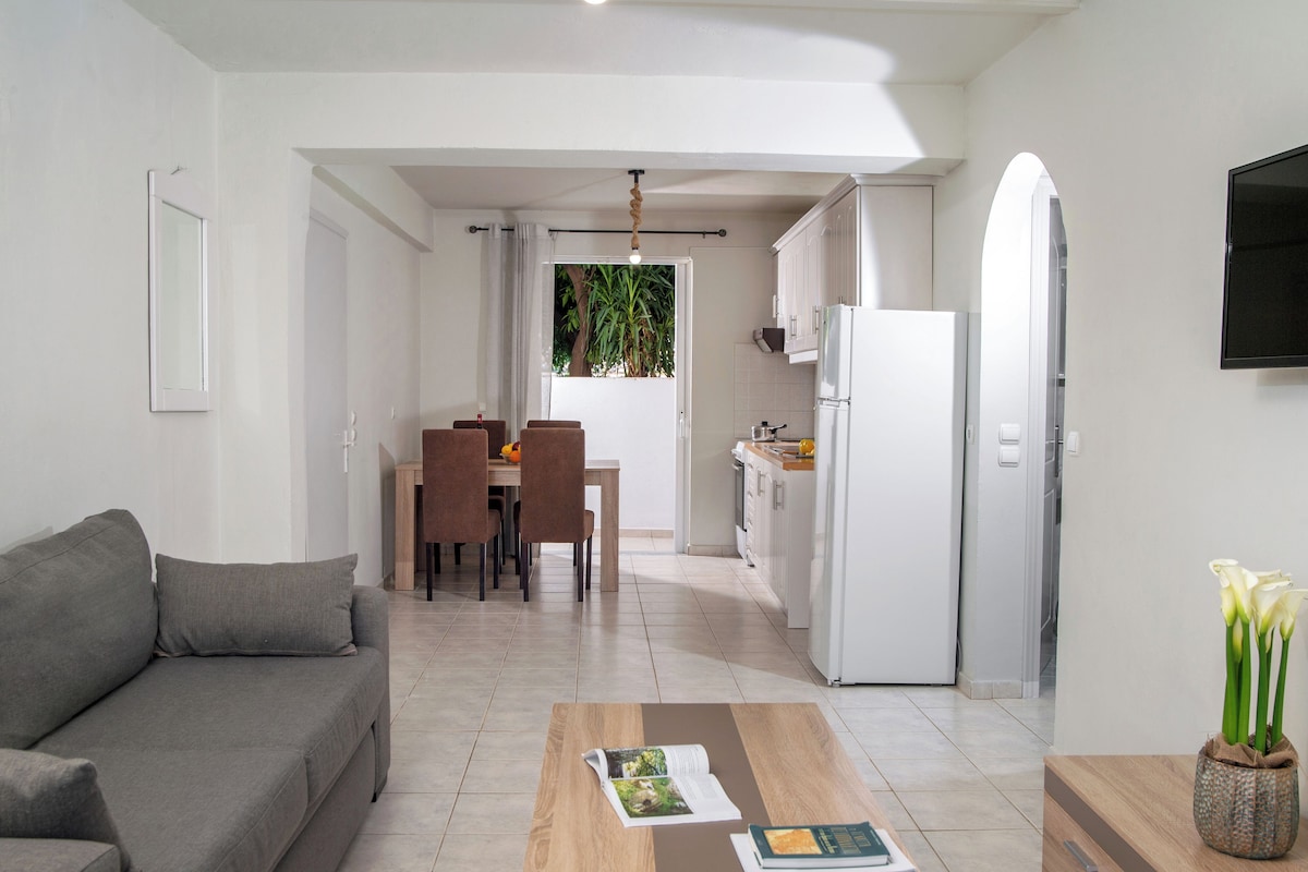 Un salon moderne comprenant un canapé, une table basse avec des magazines, un coin repas avec des chaises, une cuisine avec un réfrigérateur et un téléviseur fixé au mur.