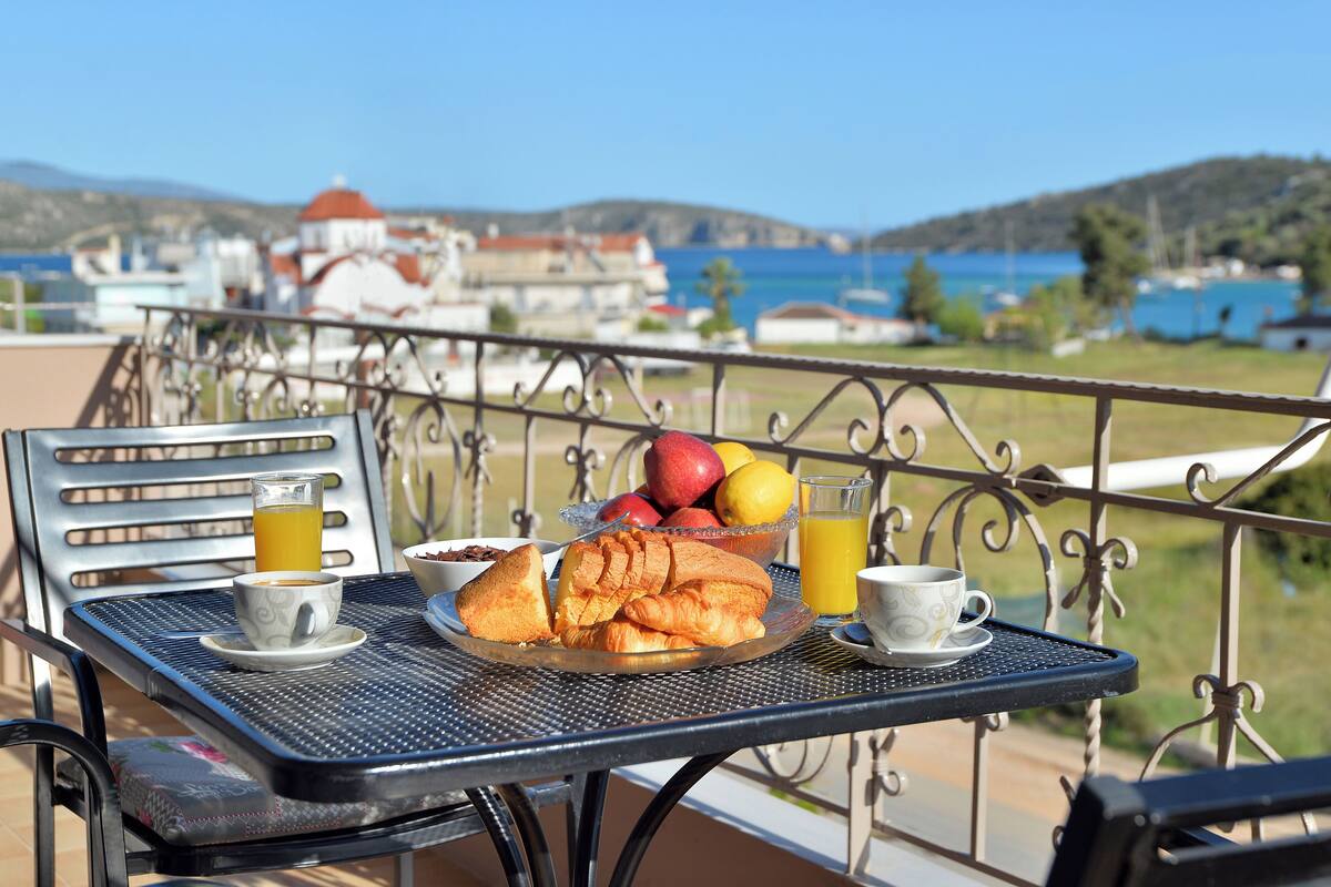 Mesa en el balcón con alimentos para el desayuno, como croissants, fruta y bebidas, con vistas a un paisaje pintoresco de una ciudad costera.