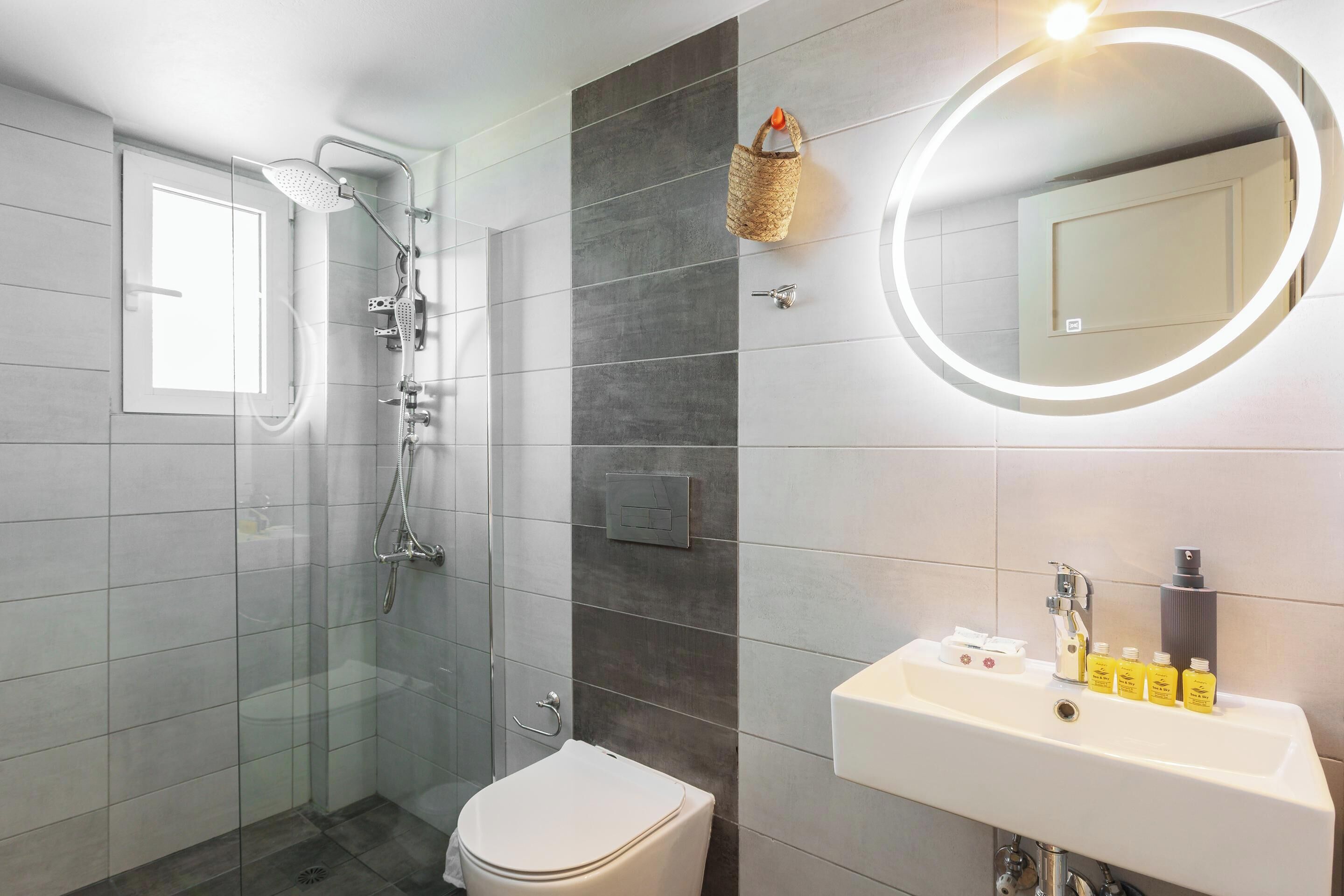 A modern bathroom with a glass-enclosed shower, a toilet, a sink with toiletries, and a wall-mounted mirror with a light.