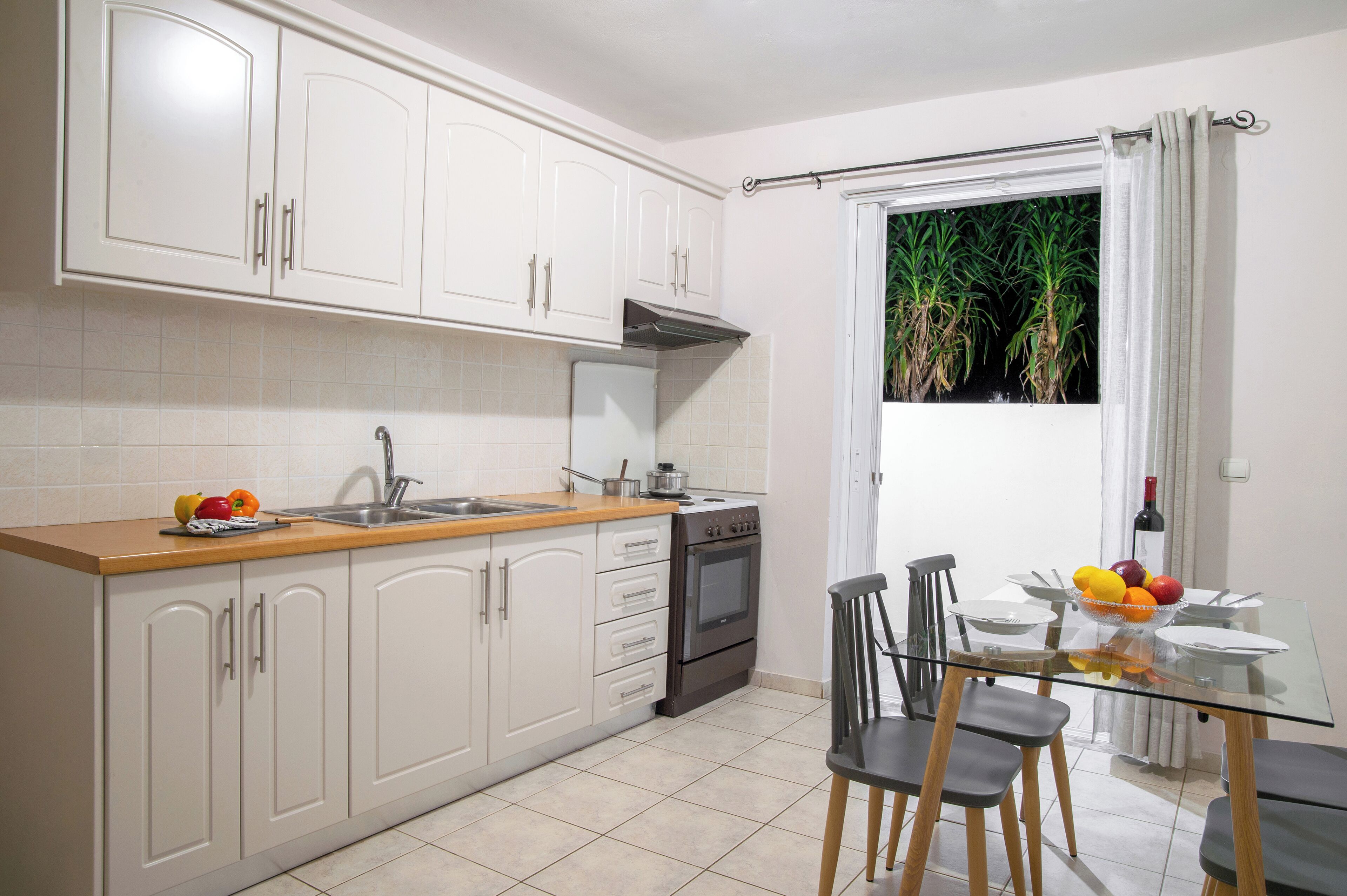 Une cuisine avec des meubles blancs, un plan de travail en bois, une cuisinière et un coin repas comprenant une table et des chaises en verre.