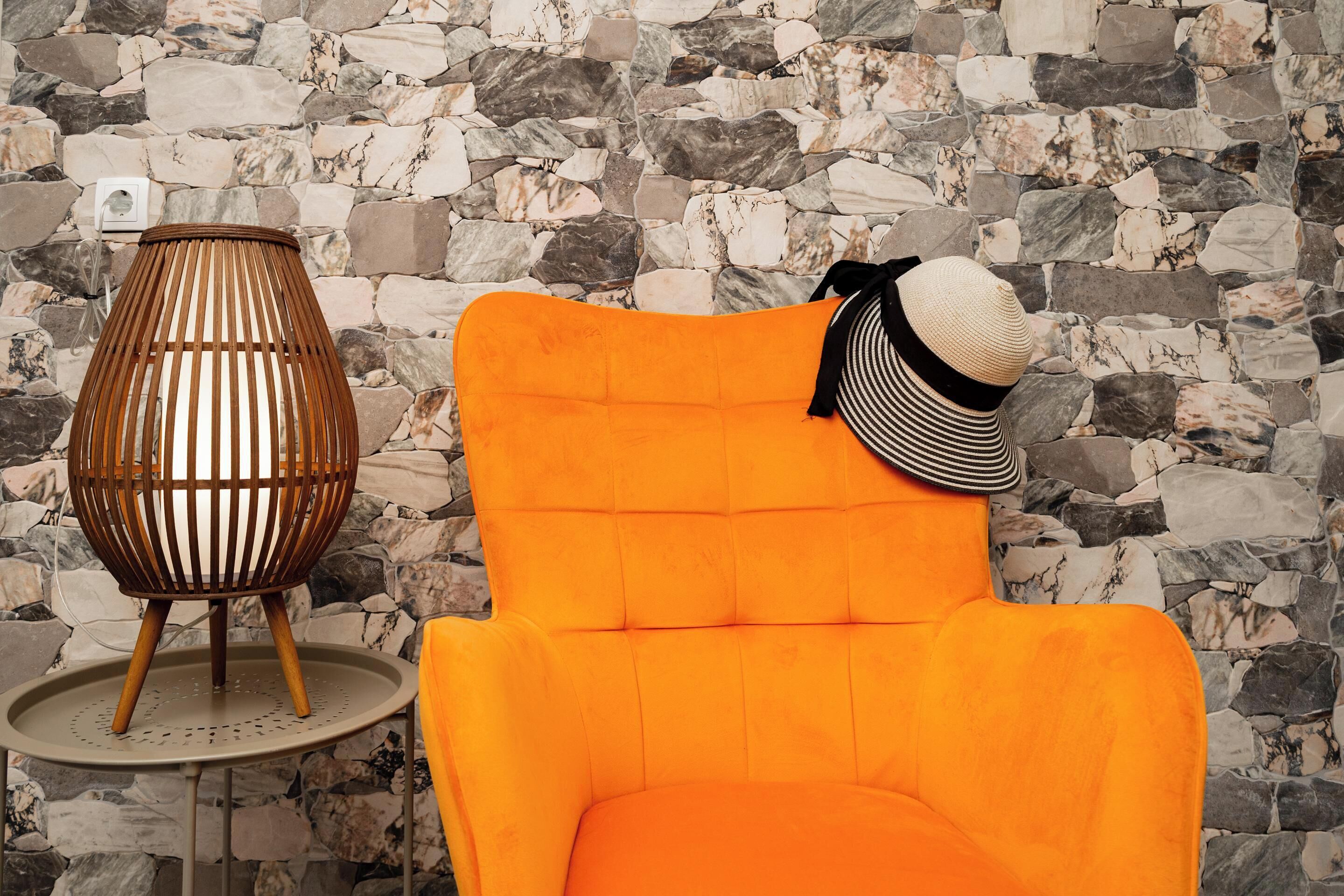 Une pièce avec un mur en pierre, une table ronde et une chaise en osier, ainsi qu’un fauteuil orange vif.