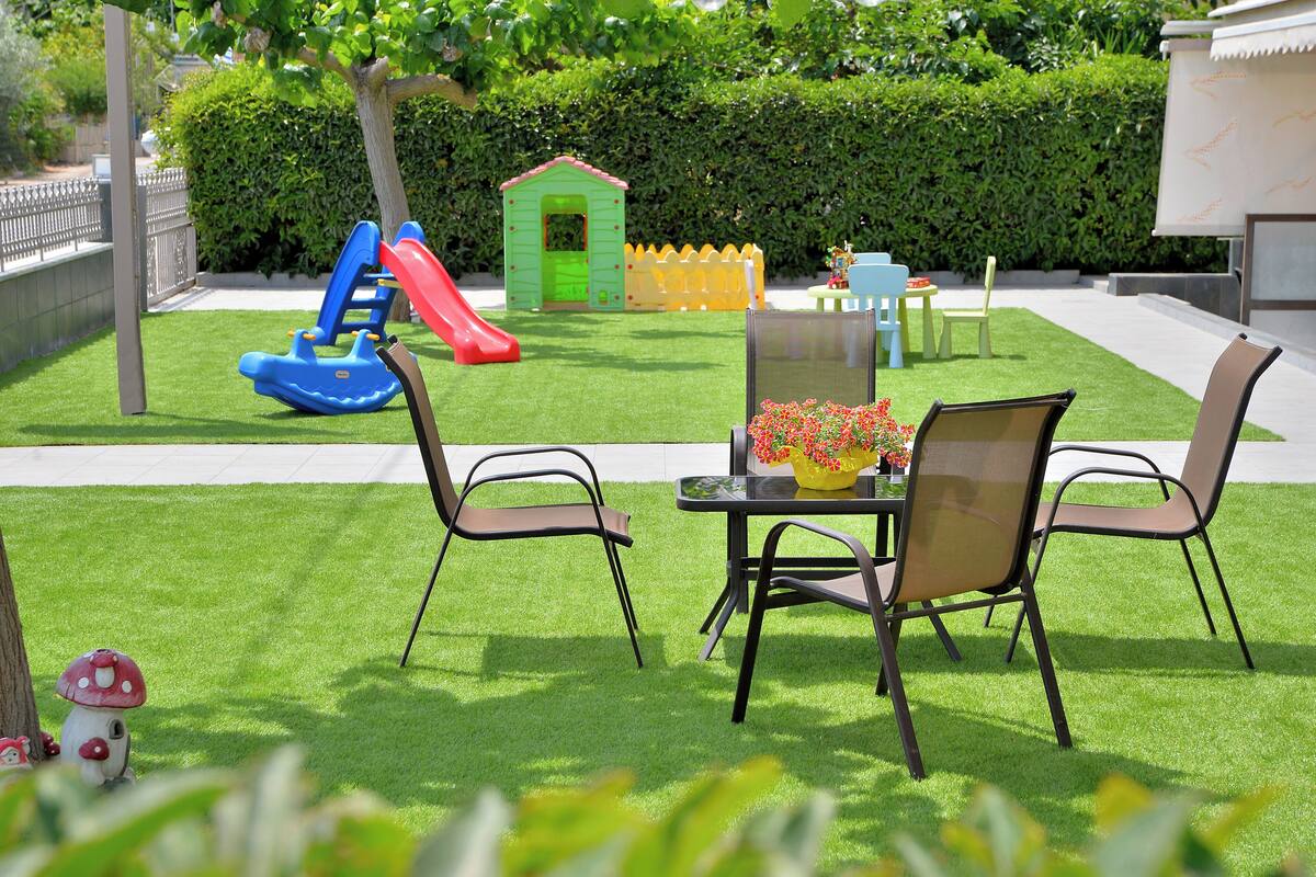 Un patio con mesa y sillas, un pequeño espacio de juegos con un tobogán, y un seto al fondo.