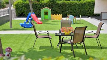 Un patio con mesa y sillas, un pequeño espacio de juegos con un tobogán, y un seto al fondo.
