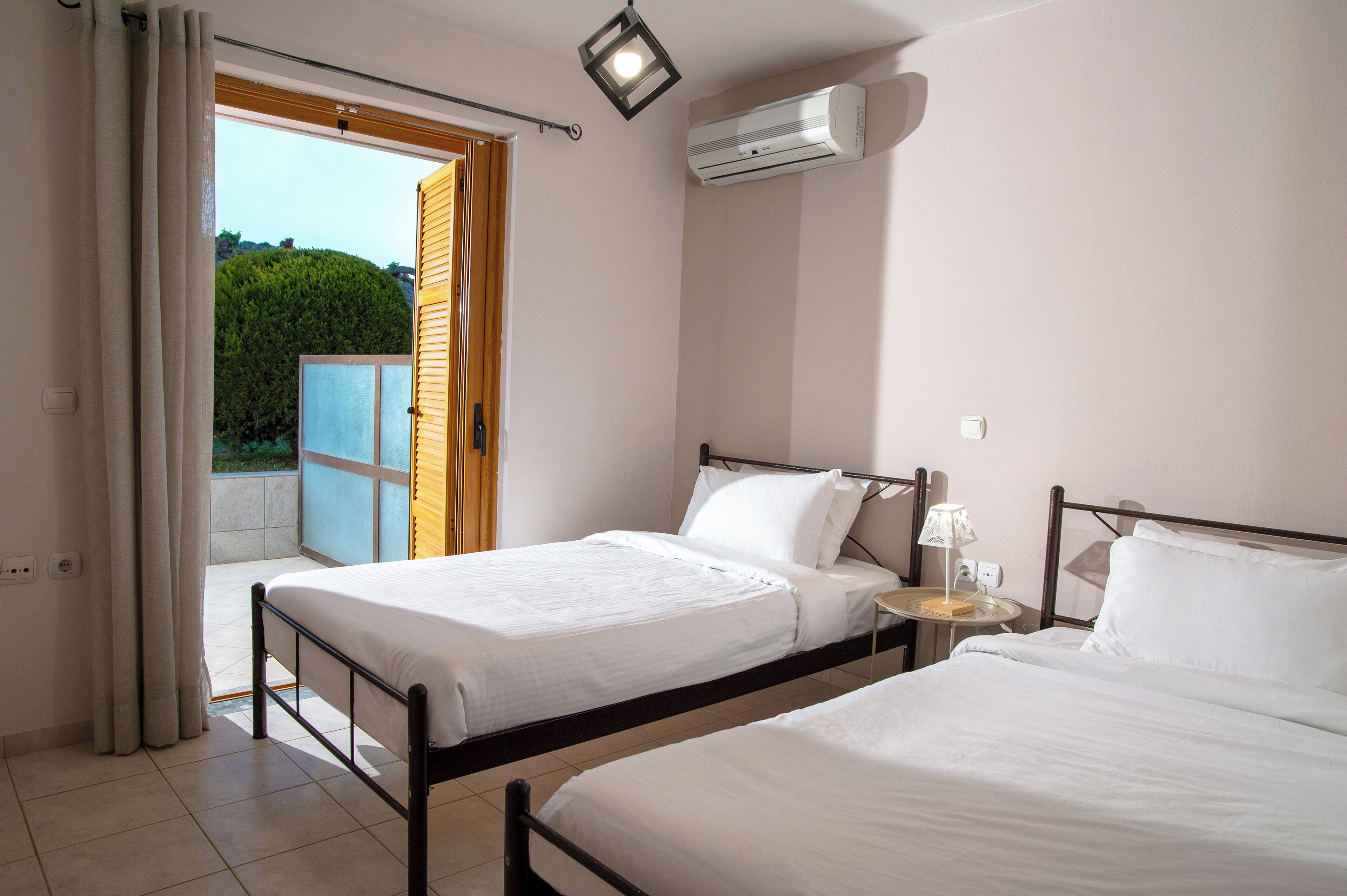 A bedroom with two beds, a small bedside table with a lamp, an air conditioning unit, and a sliding glass door leading to an outdoor area.