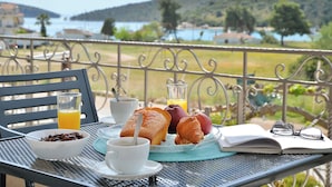 Una tavola imbandita con croissant, una ciotola di cioccolato, un bicchiere di succo d'arancia e una tazza di caffè su un balcone con vista panoramica su colline e acqua.
