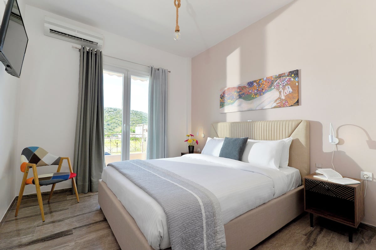 Habitación de hotel con cama, una silla, mesita de noche, televisión montada en la pared y ventana con cortinas.