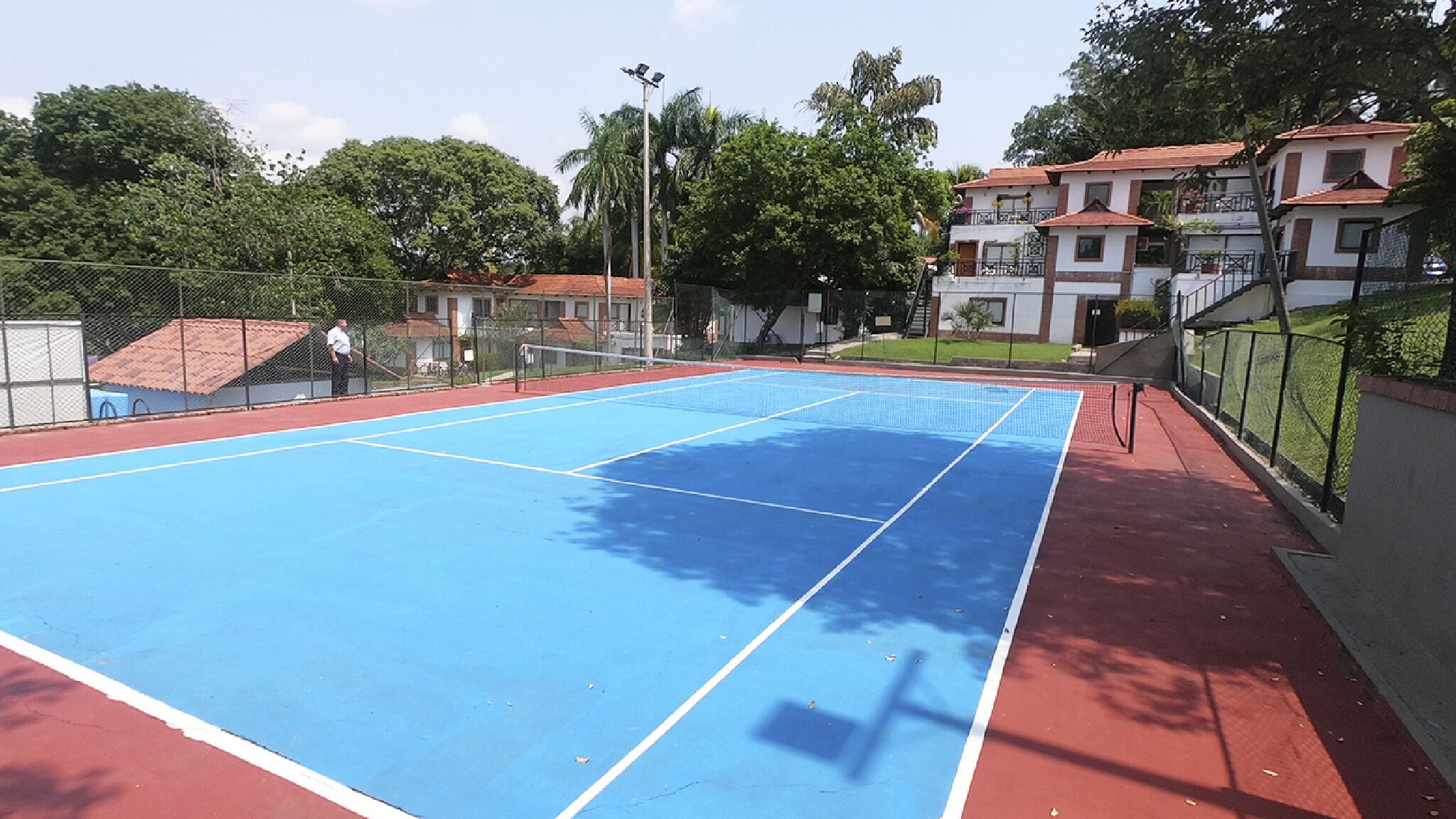 Un court de tennis extérieur doté d’une surface rouge et bleue, entouré d’une clôture et d’arbres.