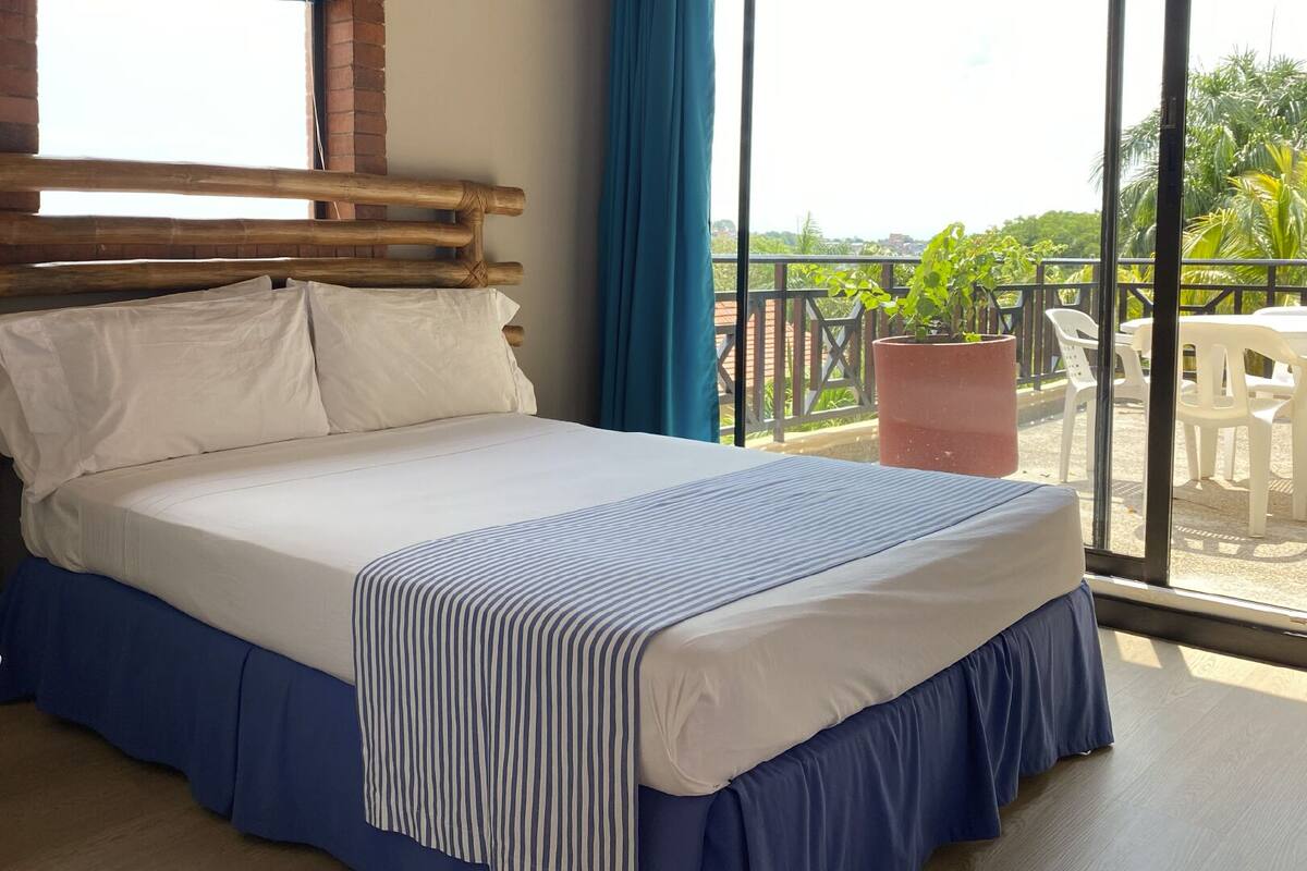 Hotelkamer met een houten hoofdeinde, een groot bed met wit en blauw beddengoed, en uitzicht op het buiten via een balkon met stoelen en een potplant.