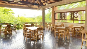 An outdoor dining area with wooden tables and chairs, surrounded by a wooden deck and large windows offering a view of lush greenery. - Hotel Girardot Resort by On Vacation (Ricaurte)