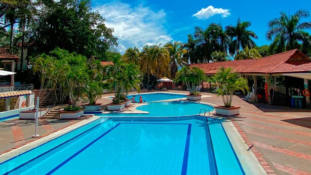 A swimming pool with a tiled deck, surrounded by palm trees and covered structures. - Hotel Girardot Resort by On Vacation (Ricaurte)