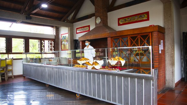 Zona de bufé con diversos alimentos exhibidos en una larga barra, una persona con uniforme de chef y un horno de ladrillo al fondo.