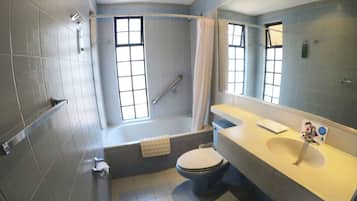 Badeværelse med badekar, toilet og håndvask. Vinduer giver naturligt lys, og der er en badeforhæng.