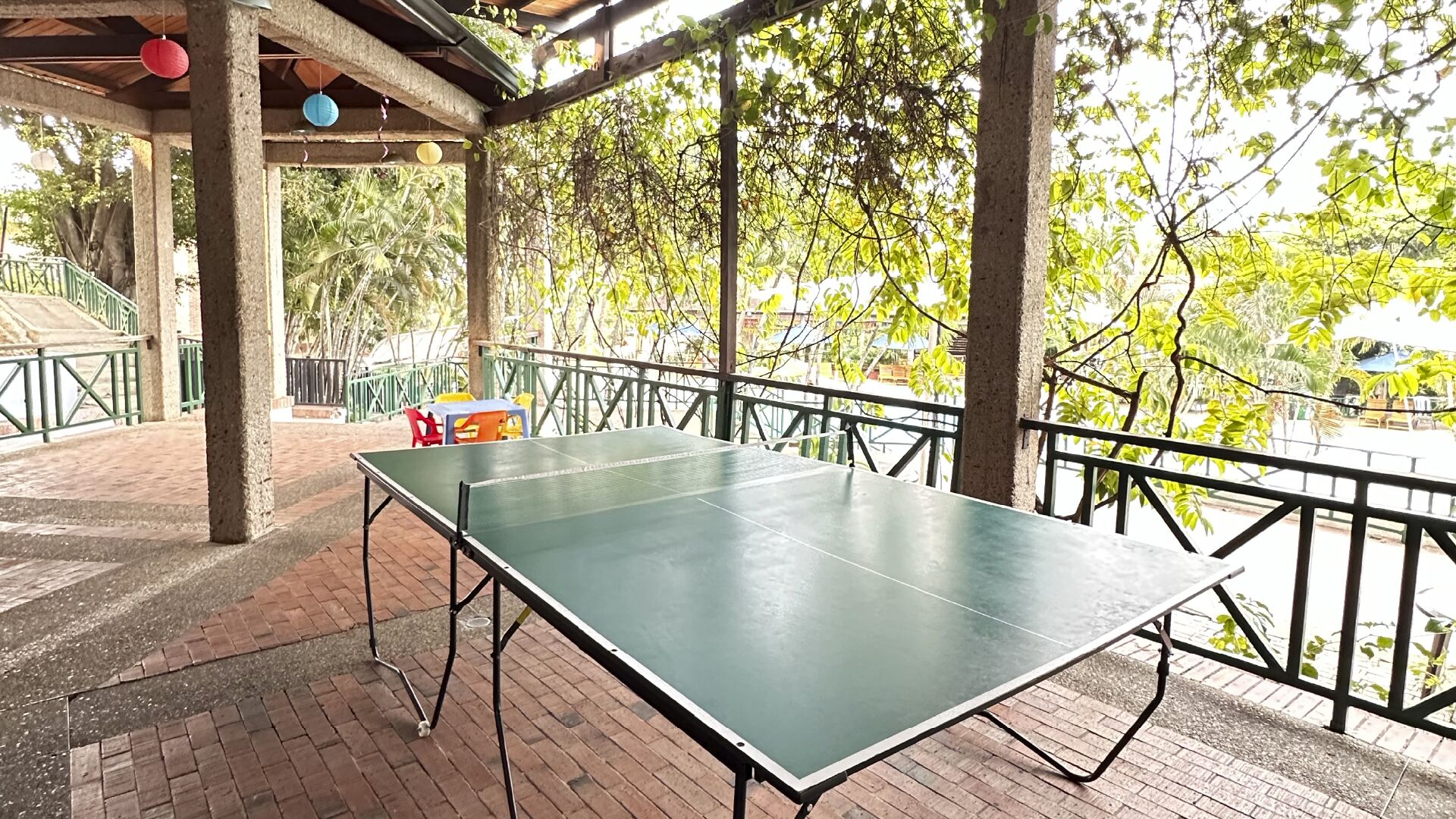An outdoor table tennis table under a covered area with a brick floor, surrounded by greenery and a metal railing.