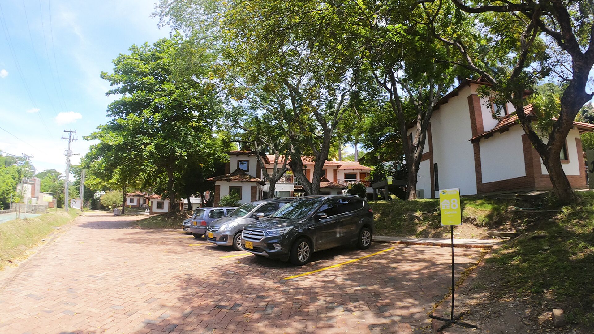 Un camino pavimentado con coches estacionados, un letrero amarillo y un edificio con techo rojo.