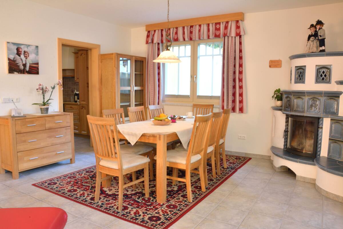 A dining area with a wooden table and chairs, a red and blue patterned rug, a kitchen with wooden cabinets, and a fireplace with decorative tiles.