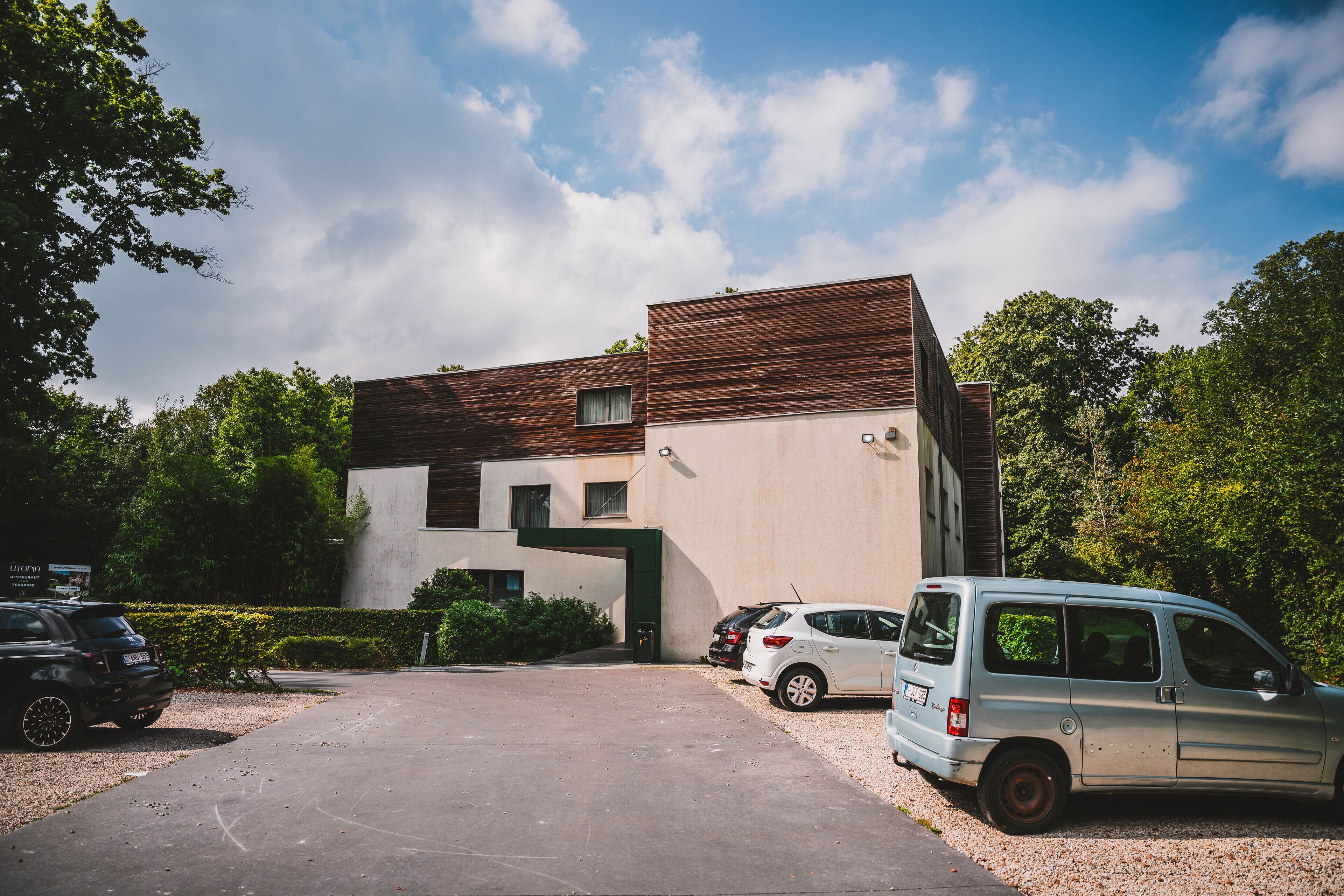 A modern building with a unique architectural design, featuring a combination of wooden and concrete materials. There are several parked cars in front of the building, including a white van and a black SUV.