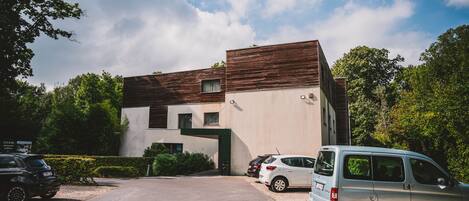 A modern building with a unique architectural design, featuring a combination of wooden and concrete materials. There are several parked cars in front of the building, including a white van and a black SUV.