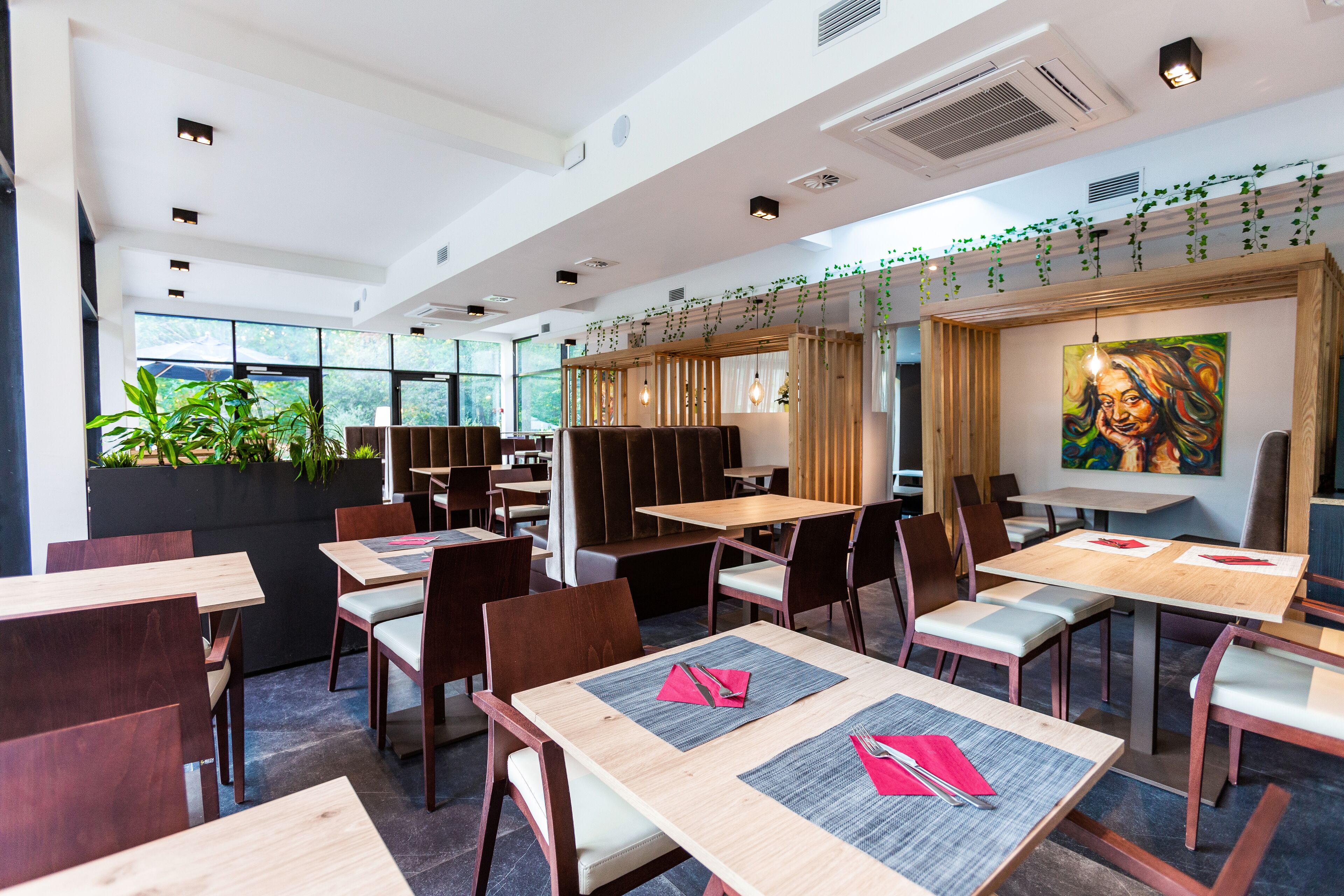 A modern restaurant with wooden tables and chairs, large windows, and indoor plants.