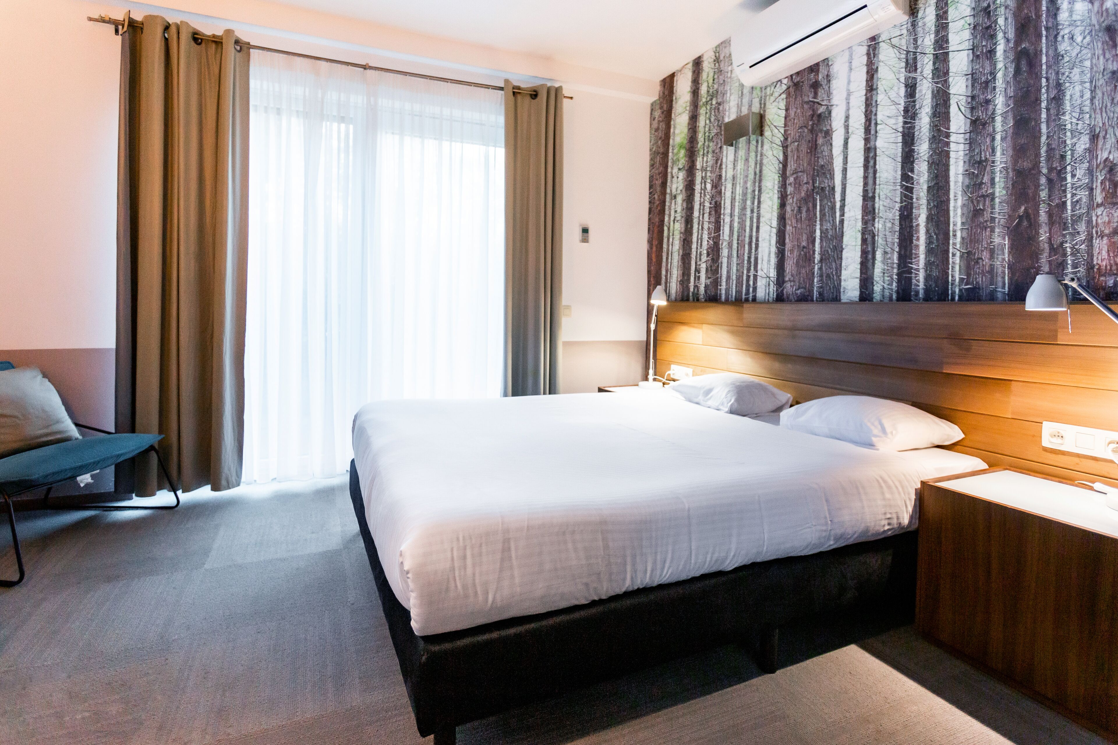 Habitación de hotel con una cama grande, una mesita de noche, un escritorio, una silla y un mural en la pared con tema de bosque.