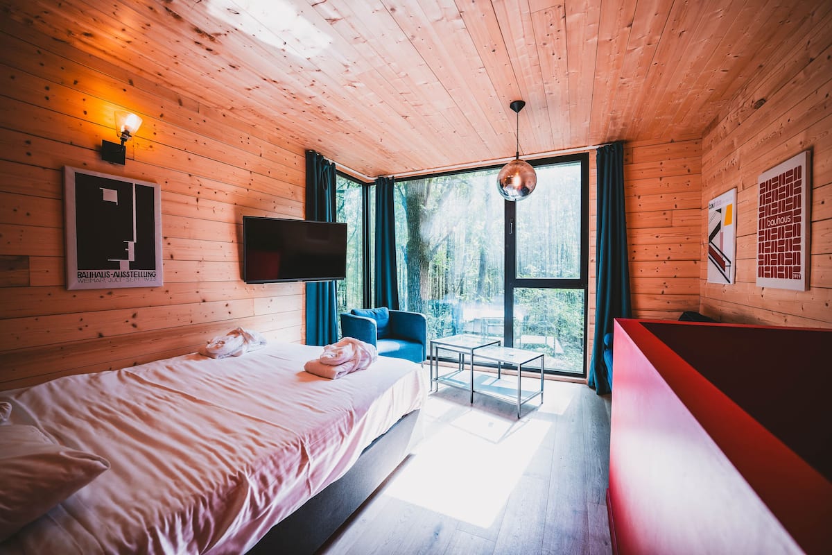Une pièce de cabane en bois comprenant un lit, un téléviseur, une chaise bleue, une petite table et un banc rouge.