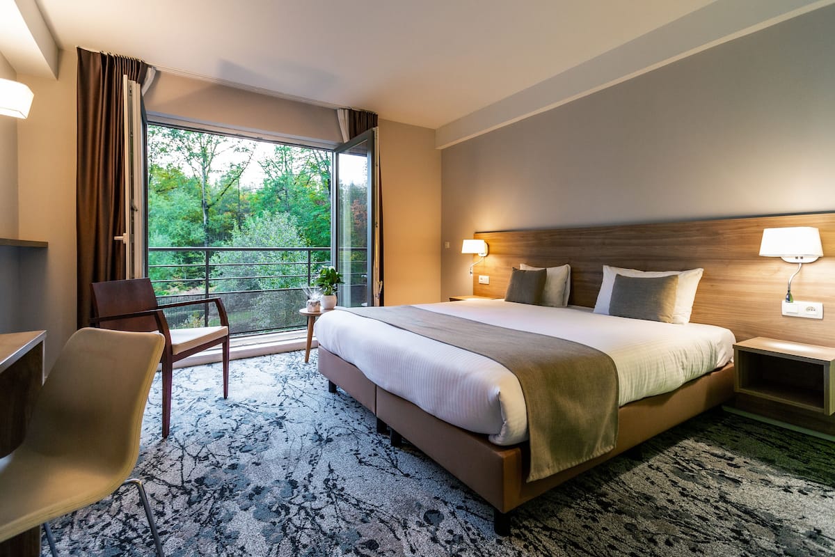 Une chambre d’hôtel avec un grand lit, un bureau, une chaise, un balcon donnant sur la verdure et deux lampes de chevet.