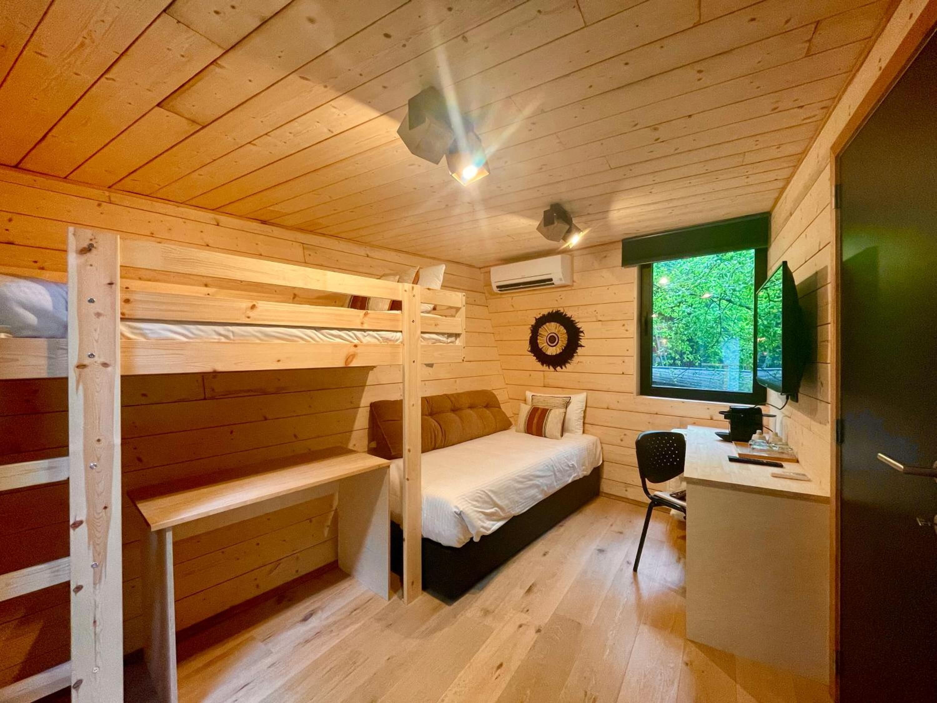 A compact room with wooden walls and floors, bunk beds, a desk with a chair, a television, and a window with a view of greenery.