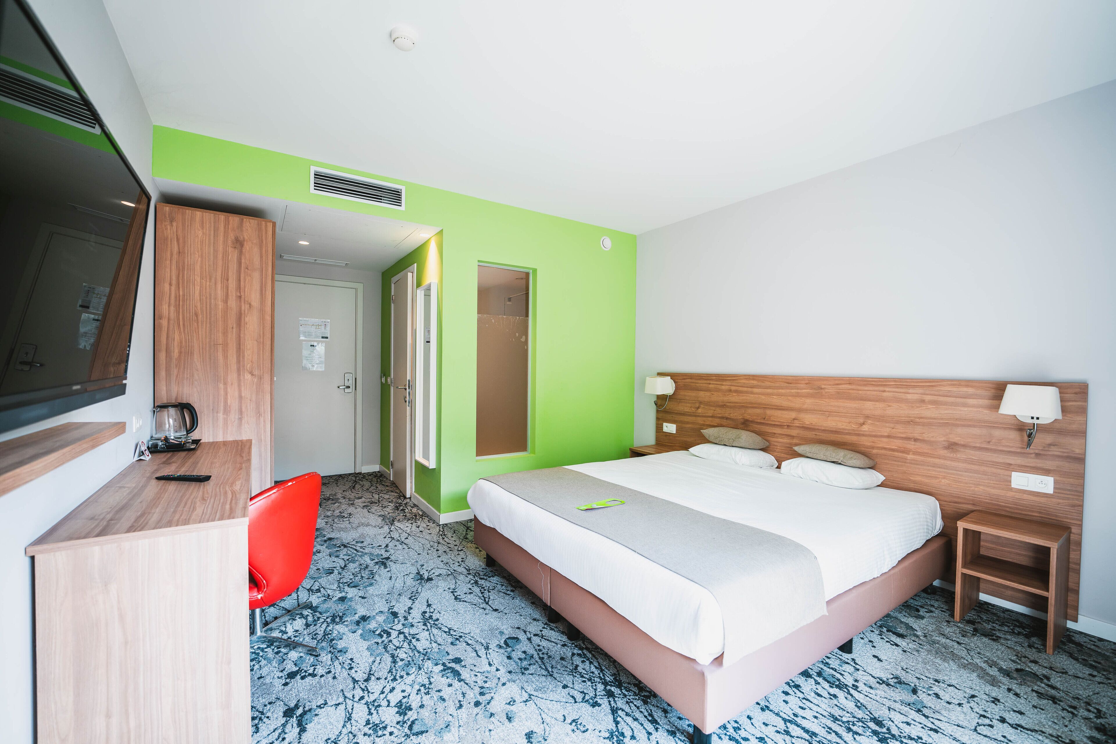 Una habitación de hotel con una cama, un escritorio, una silla roja, un cabecero de madera y una mesita de noche pequeña.