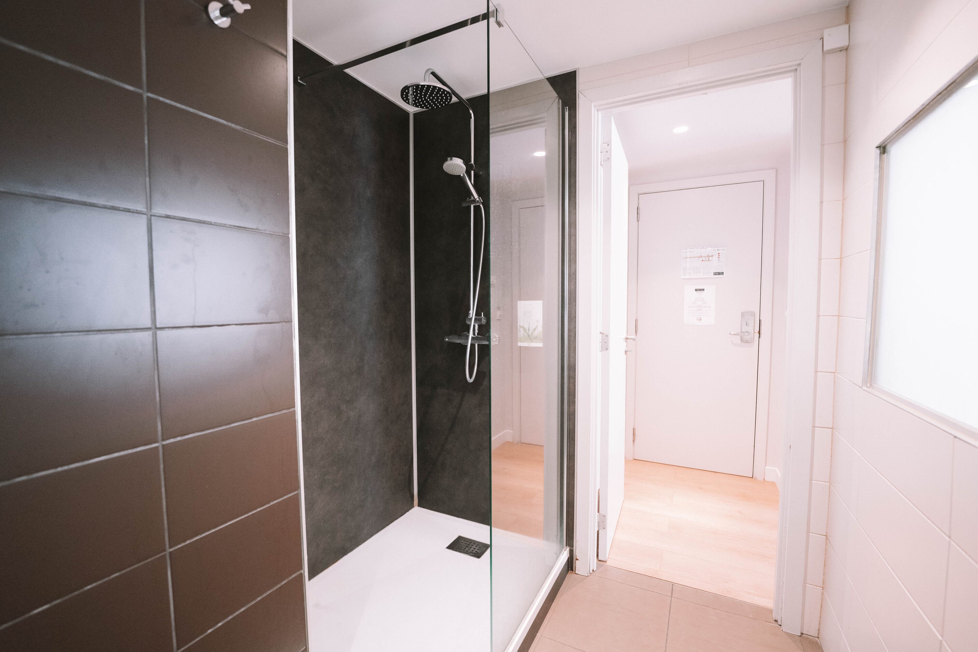A modern bathroom with a glass-enclosed shower, dark tiled walls, and a visible door leading to another room.