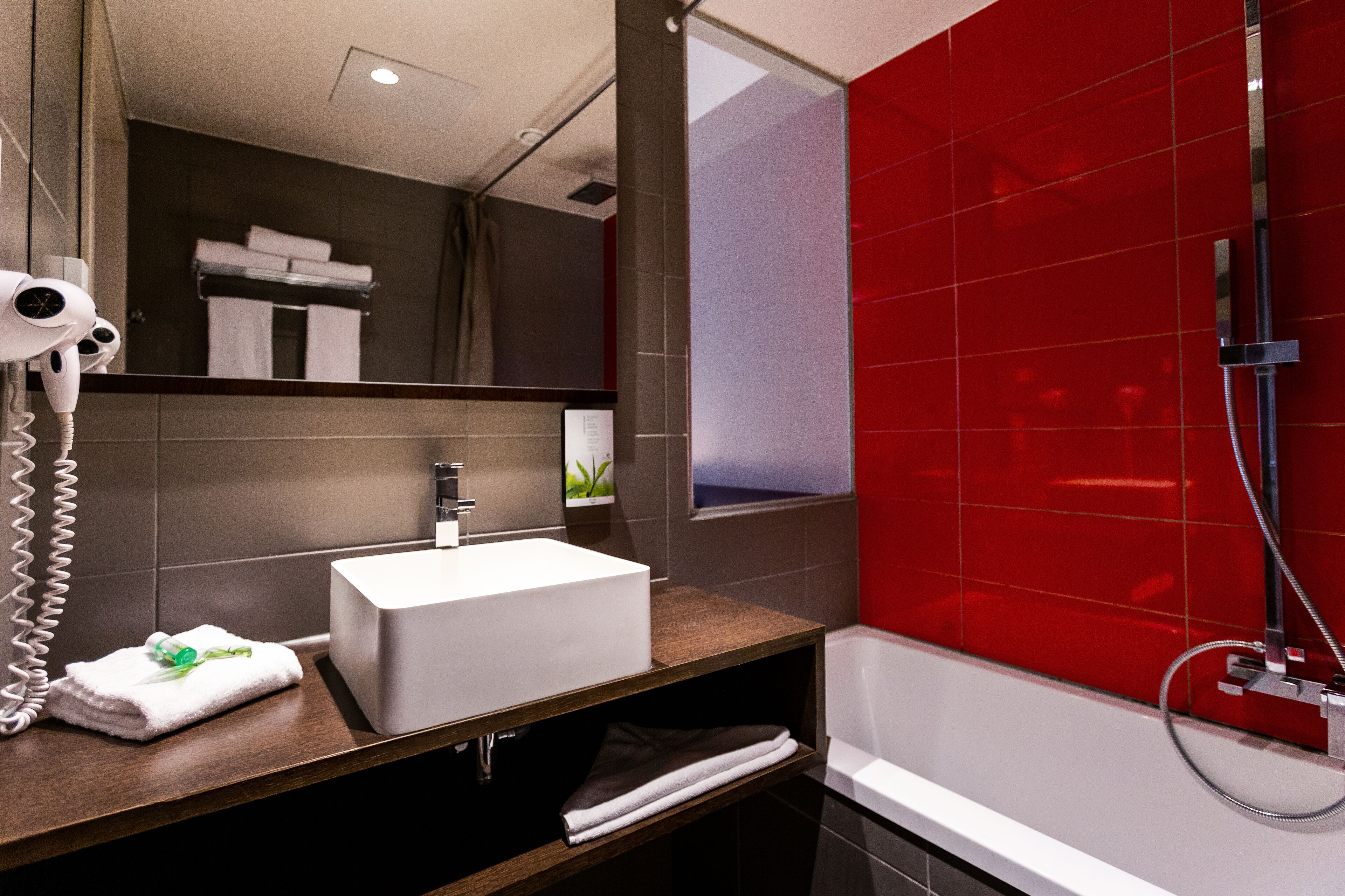 Un baño moderno con un lavamanos blanco, un secador de pelo y una zona de ducha con azulejos rojos.
