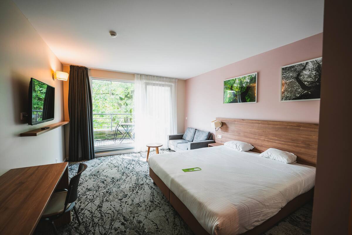 Une chambre d’hôtel avec un grand lit, un bureau, une chaise, une télévision et un balcon donnant sur un espace verdoyant.