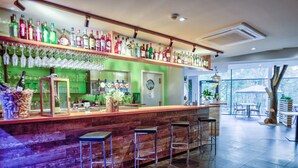 Zona de bar con mostrador de madera, varios taburetes y estanterías repletas de botellas de licor y vasos.