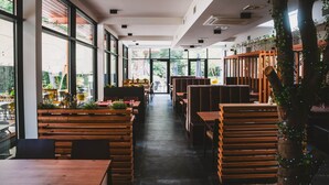 Interior moderno de restaurante con bancos y mesas de madera, ventanas amplias y plantas de interior.