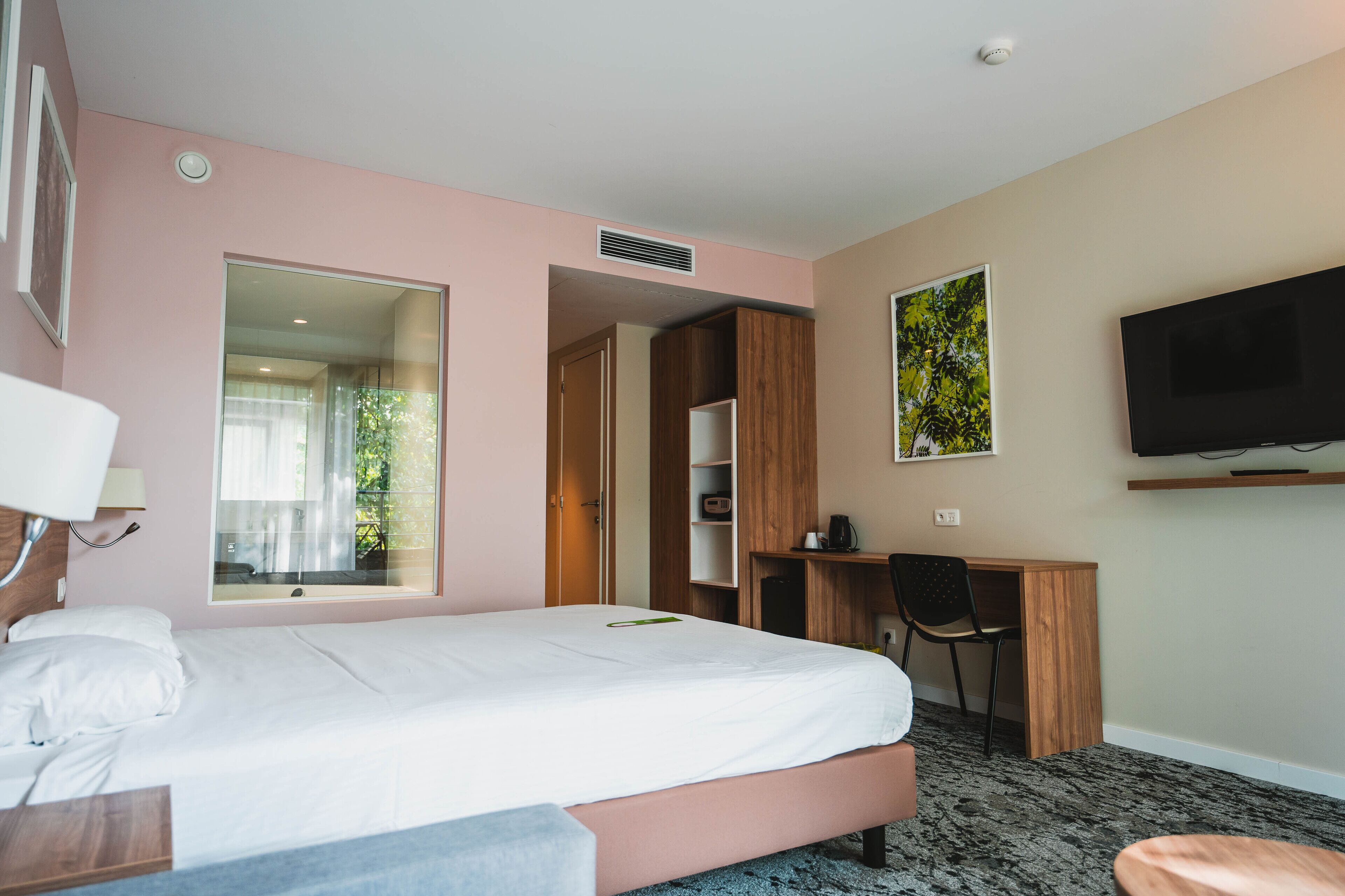 A hotel room with a bed, a desk, a chair, a TV, and a window with a view of greenery.