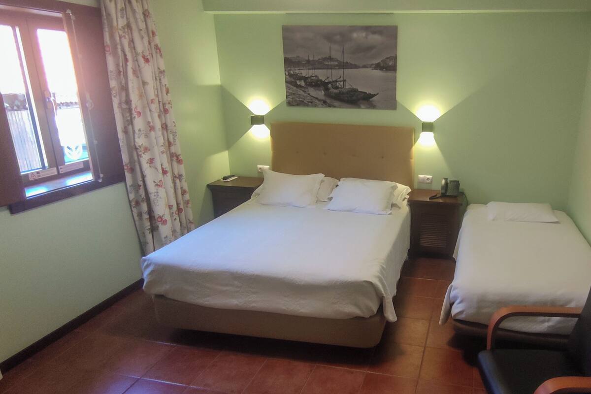 Un dormitorio con cama, mesitas de noche, una silla, una ventana con cortinas y un cuadro colgado en la pared.