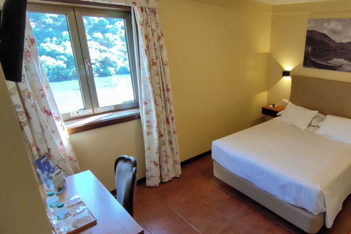 Una habitación de hotel con una cama, mesitas de noche, un escritorio con una silla, una ventana con cortinas y una lámpara de pared.