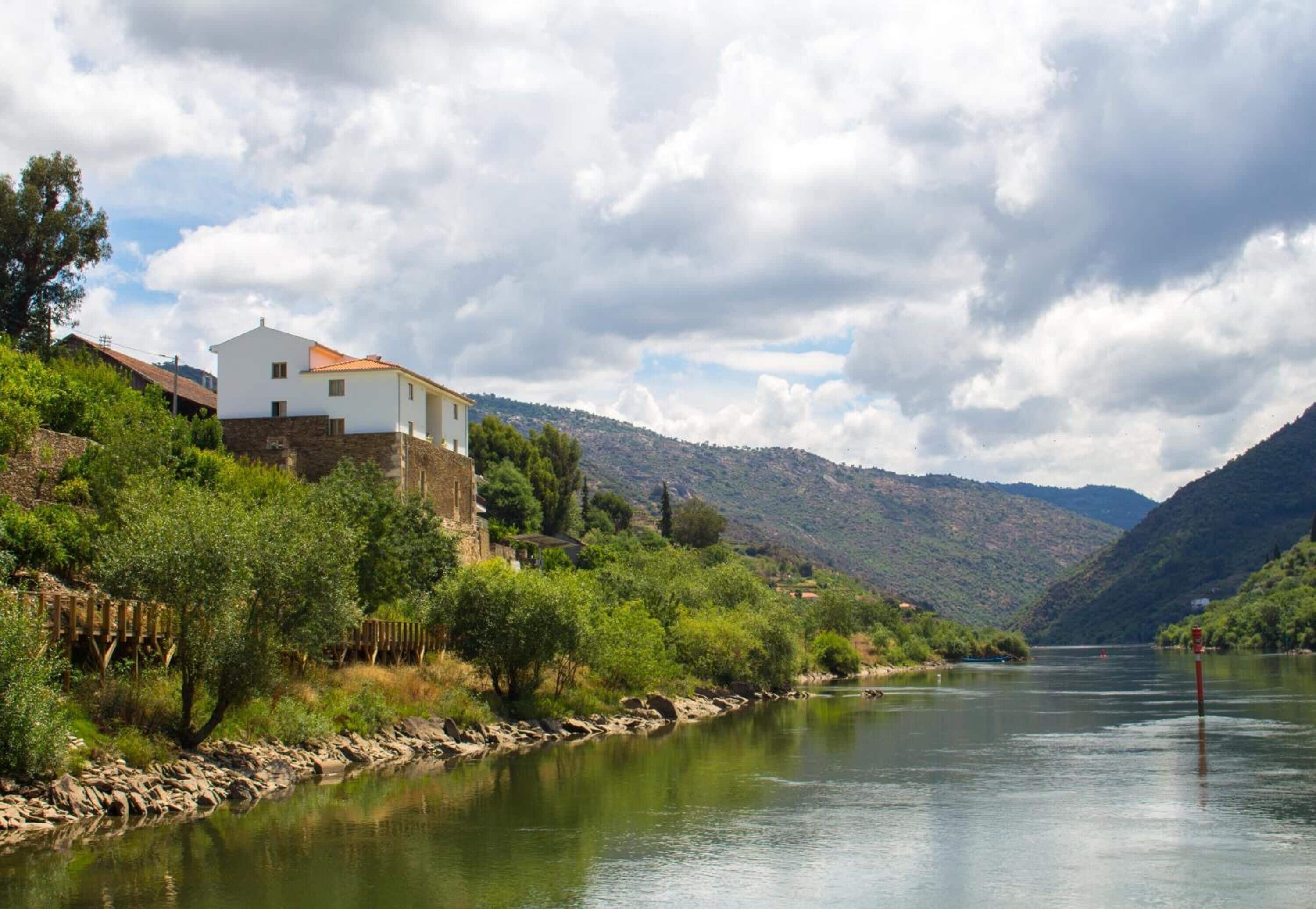 A white building with a red roof near a river, surrounded by greenery and hills.