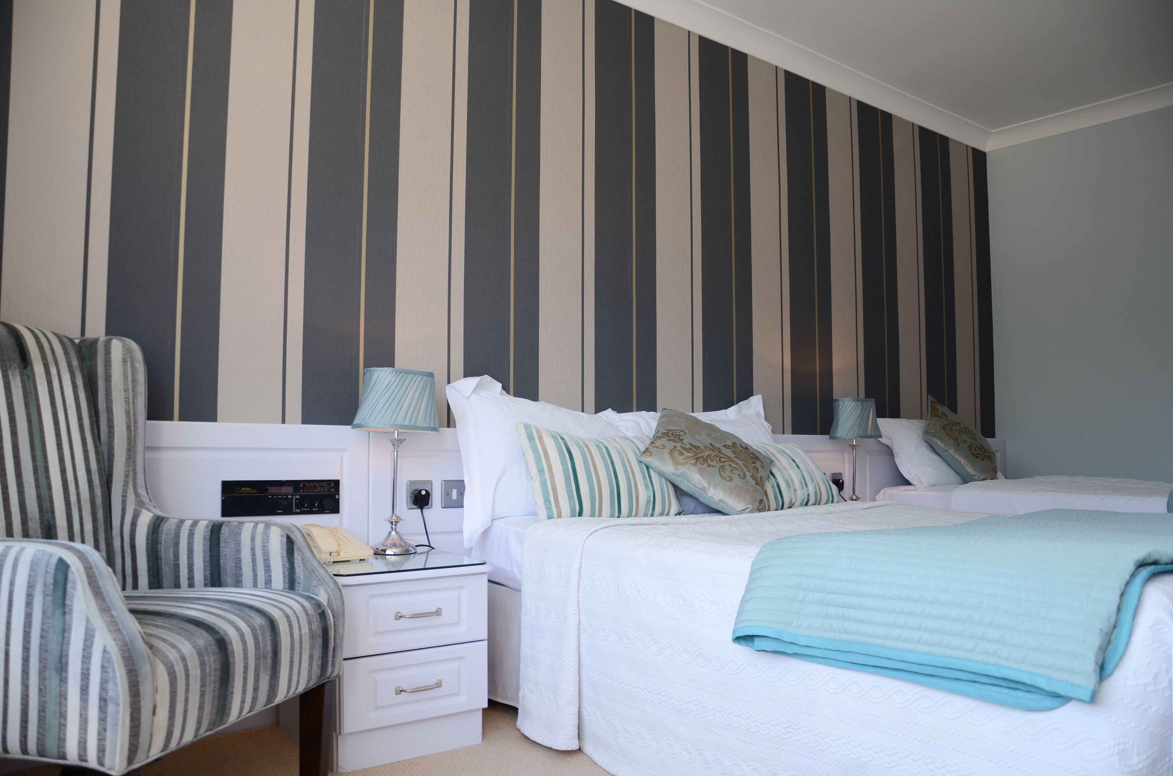 A hotel room with a striped wall, a bed with white bedding, a striped armchair, a bedside table with a lamp, and a telephone.