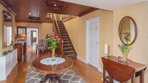 Ein geräumiges Foyer mit einer Holztreppe, einem runden Holztisch mit Spitzen-Unterlage und einem Spiegel mit Blumen.